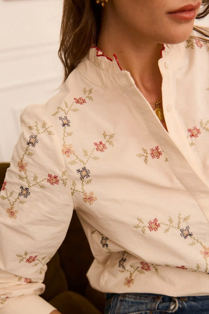 A close-up of a woman wearing a cream-colored blouse with delicate multicolor floral embroidery, paired with blue jeans.