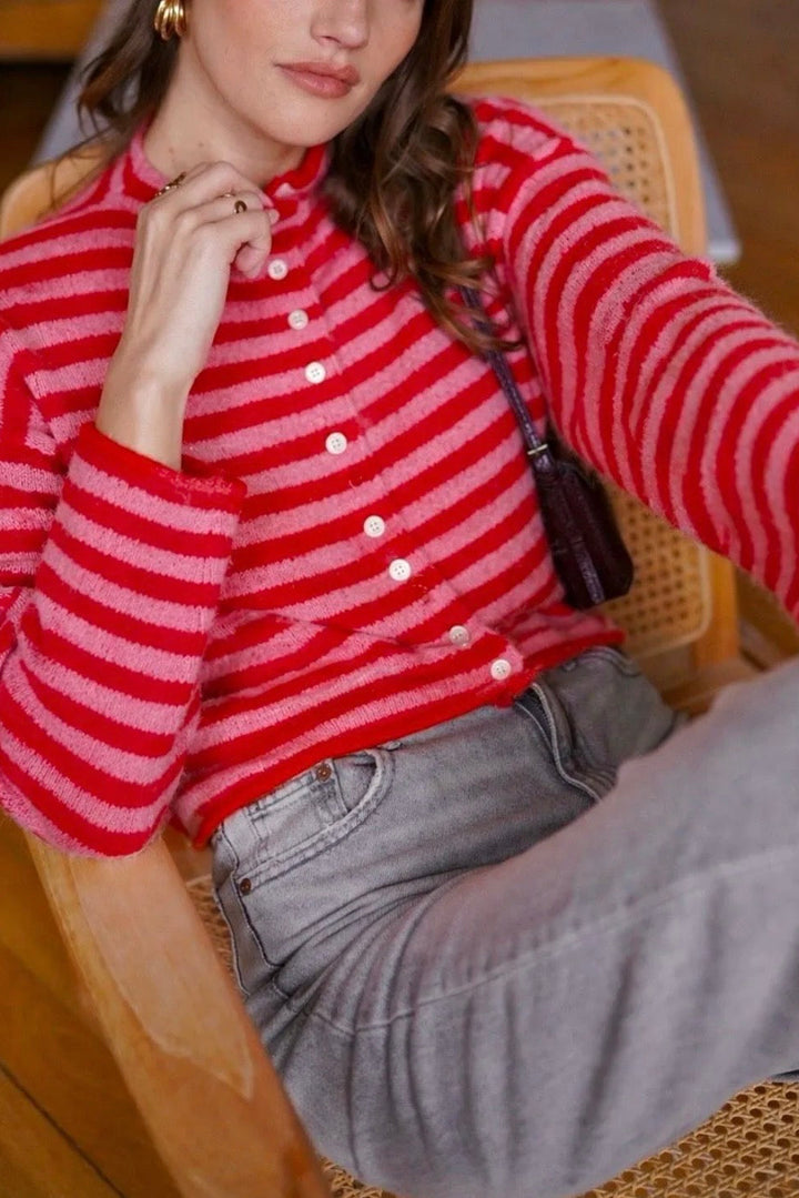 A woman seated in a wooden chair wearing a fuzzy red and pink striped button-front cardigan, light gray jeans, and gold hoop earrings in an indoor setting with a wood floor.