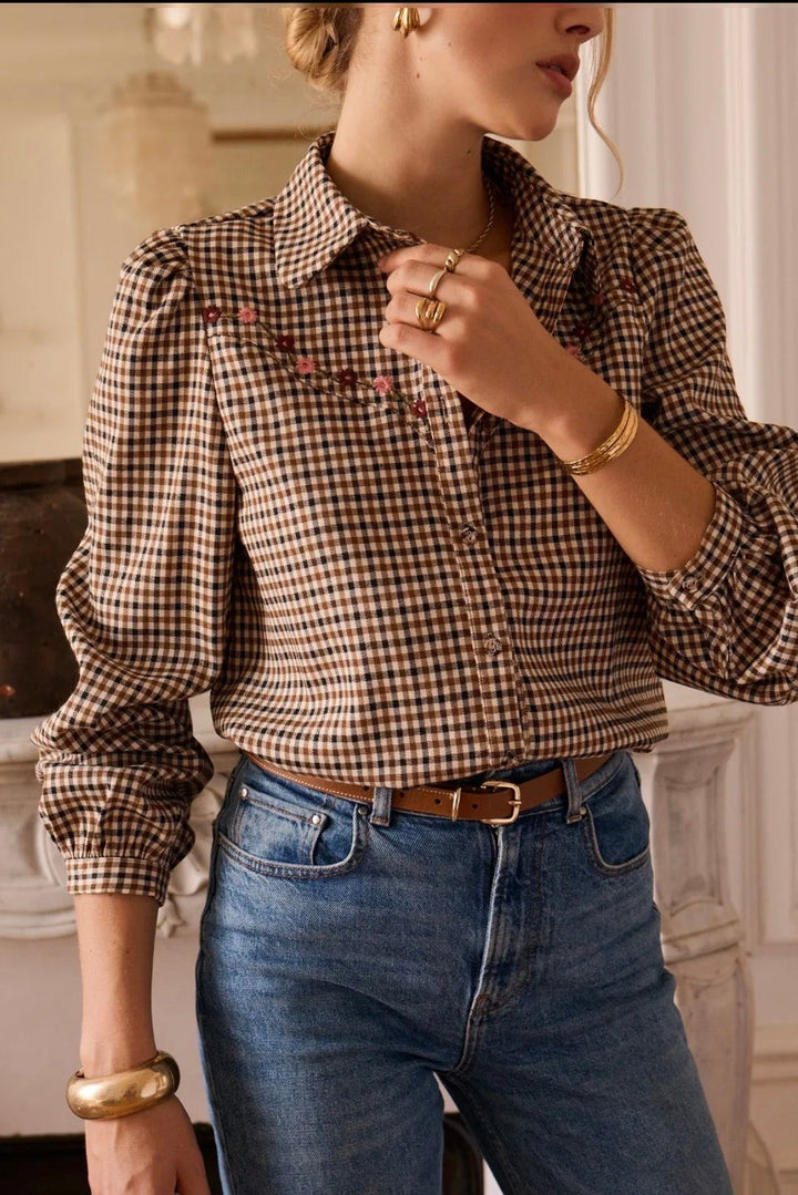 A woman wearing a brown and beige gingham long-sleeve button-up shirt with delicate floral embroidery across the chest, tucked into blue jeans with a brown belt.