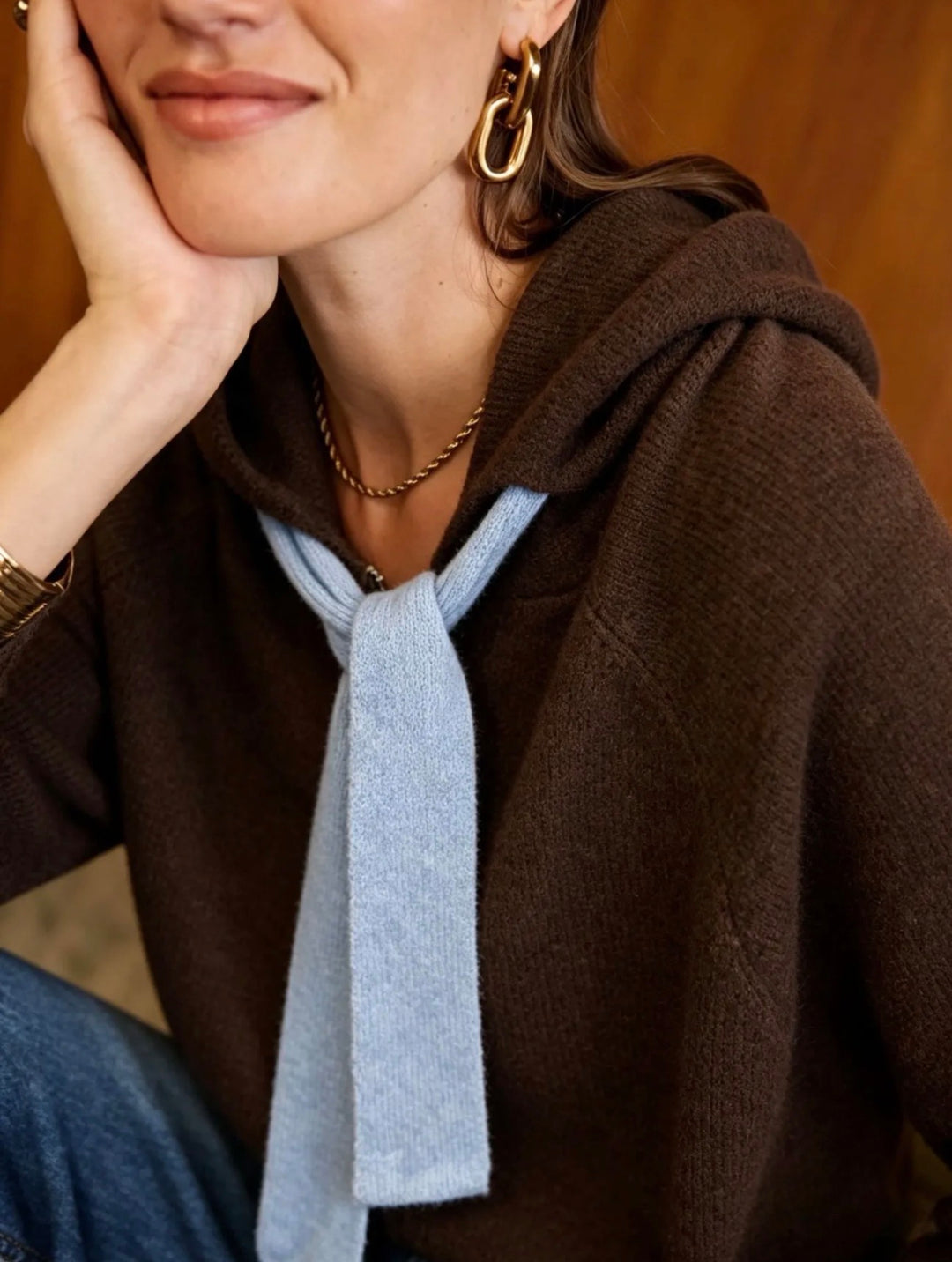 A close-up lifestyle shot of a woman wearing a dark brown knit hoodie over a light blue knit tie-neck sweater, accessorized with gold hoop earrings and a delicate gold chain necklace.