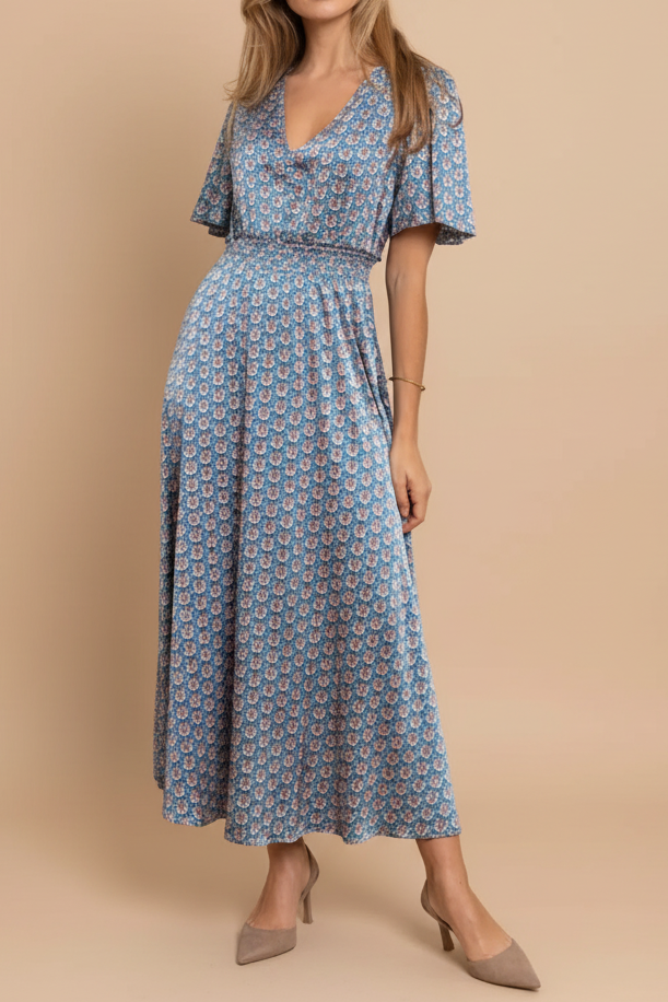 Woman in a blue patterned dress standing in a room with wooden furniture and white walls.