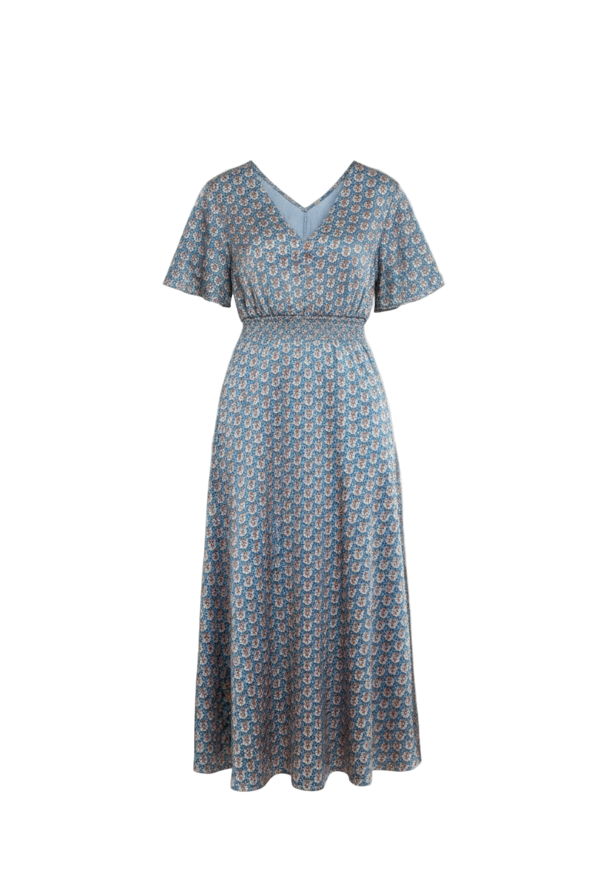 Woman in a blue patterned dress standing in a room with wooden furniture and white walls.
