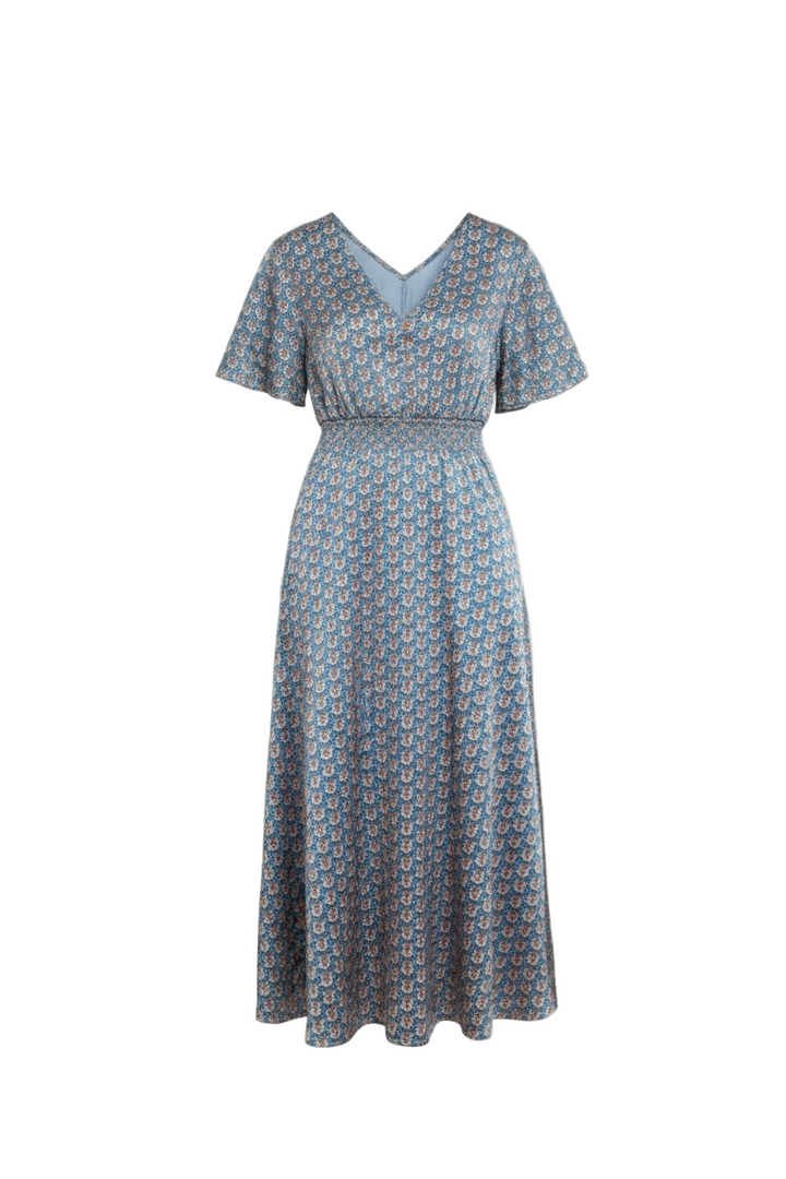 Woman in a blue patterned dress standing in a room with wooden furniture and white walls.