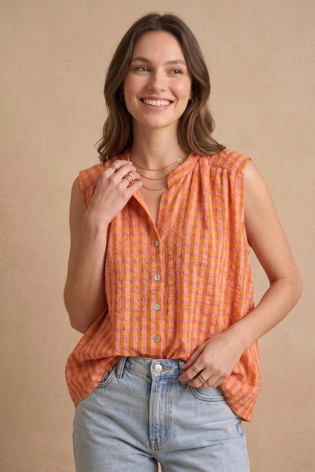 A woman wearing a sleeveless orange and white gingham button-up top with light-wash jeans, standing against a textured beige studio backdrop.