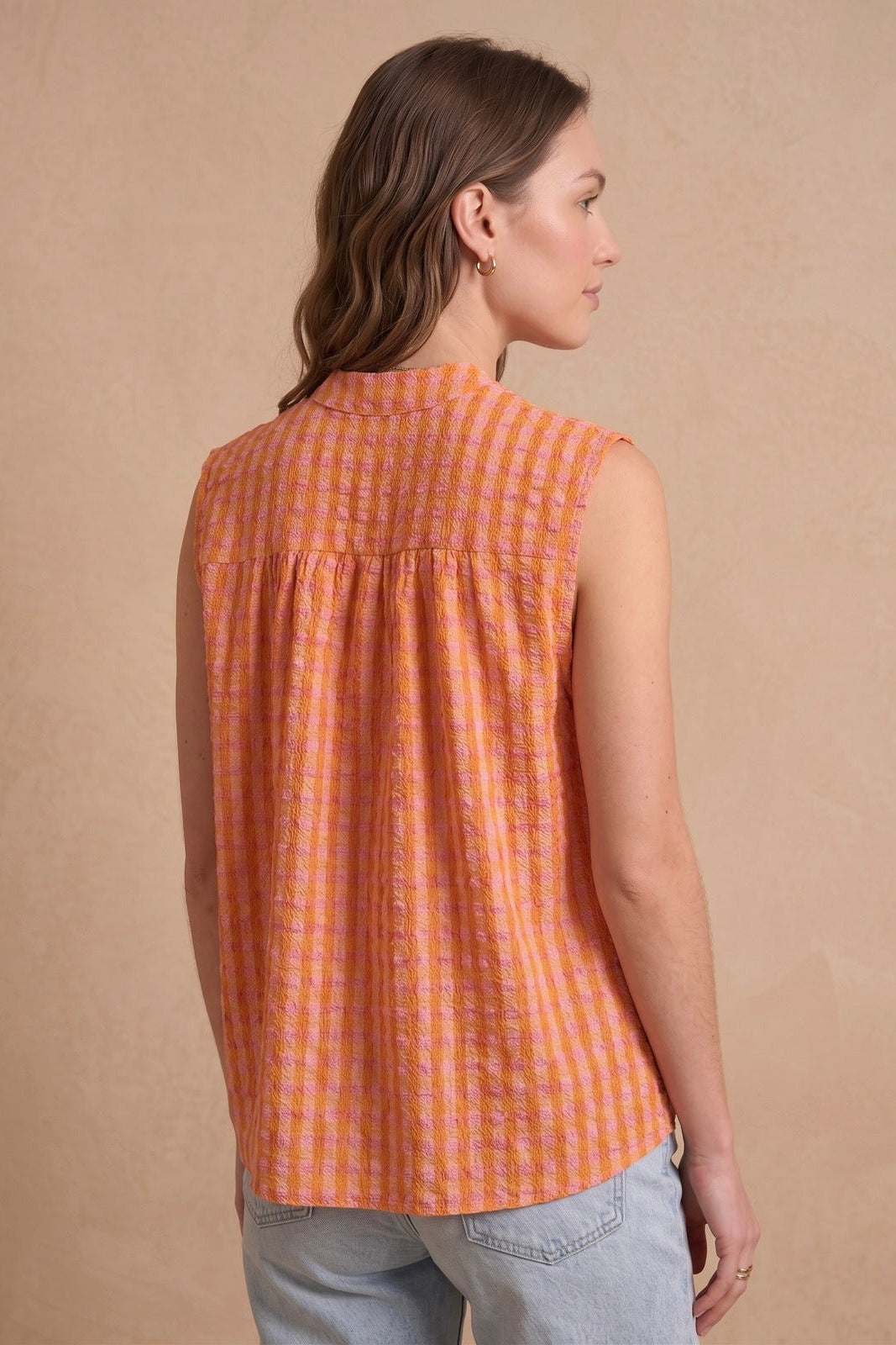 A woman shown from the back wearing a sleeveless orange and white checkered blouse with a high collar, paired with light blue jeans, against a textured beige background.