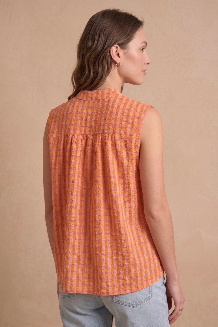 A woman shown from the back wearing a sleeveless orange and white checkered blouse with a high collar, paired with light blue jeans, against a textured beige background.