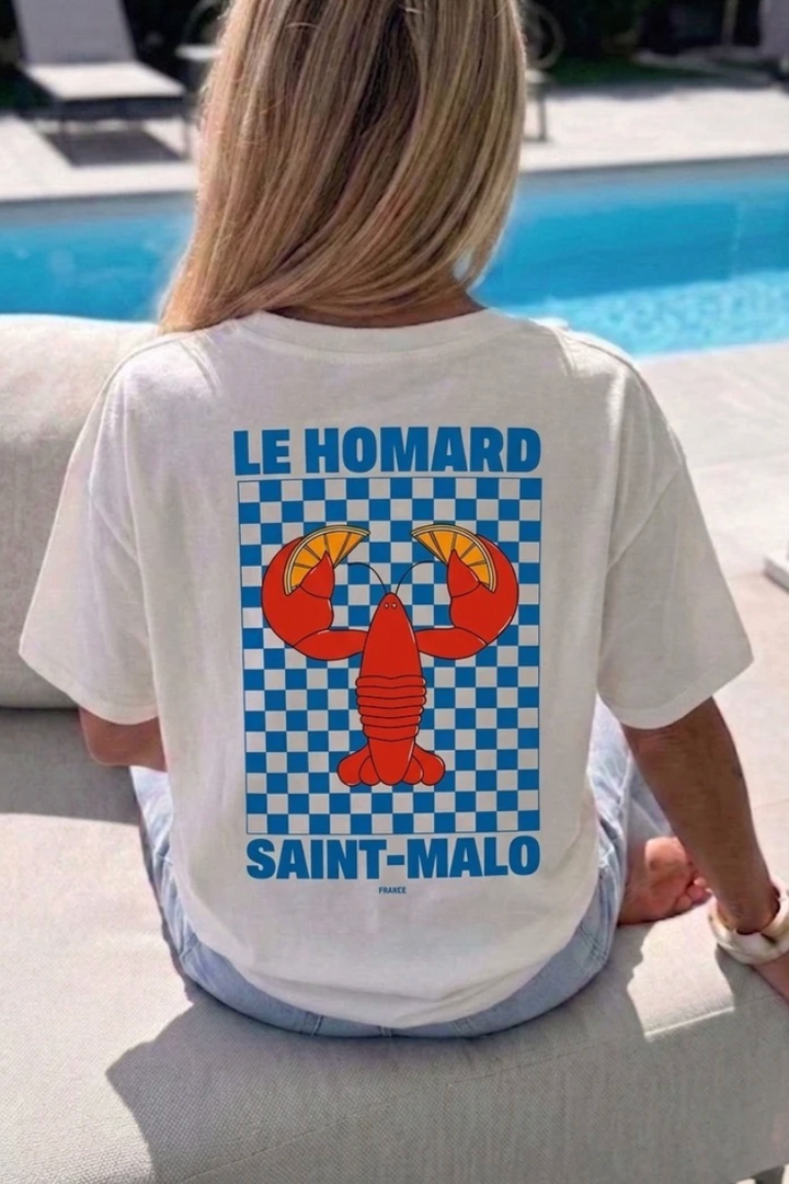 A woman sitting outdoors by a swimming pool wearing a white graphic T-shirt. The back of the shirt features a large blue and white checkered square with a red lobster graphic and the text 'LE HOMARD SAINT-MALO' in blue.