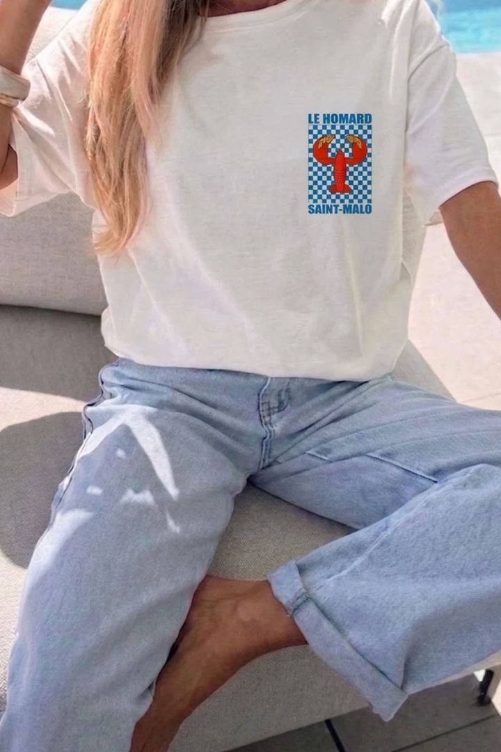 A person sitting outdoors wearing a white short-sleeve T-shirt with a blue and red lobster graphic on the chest, paired with light blue jeans.