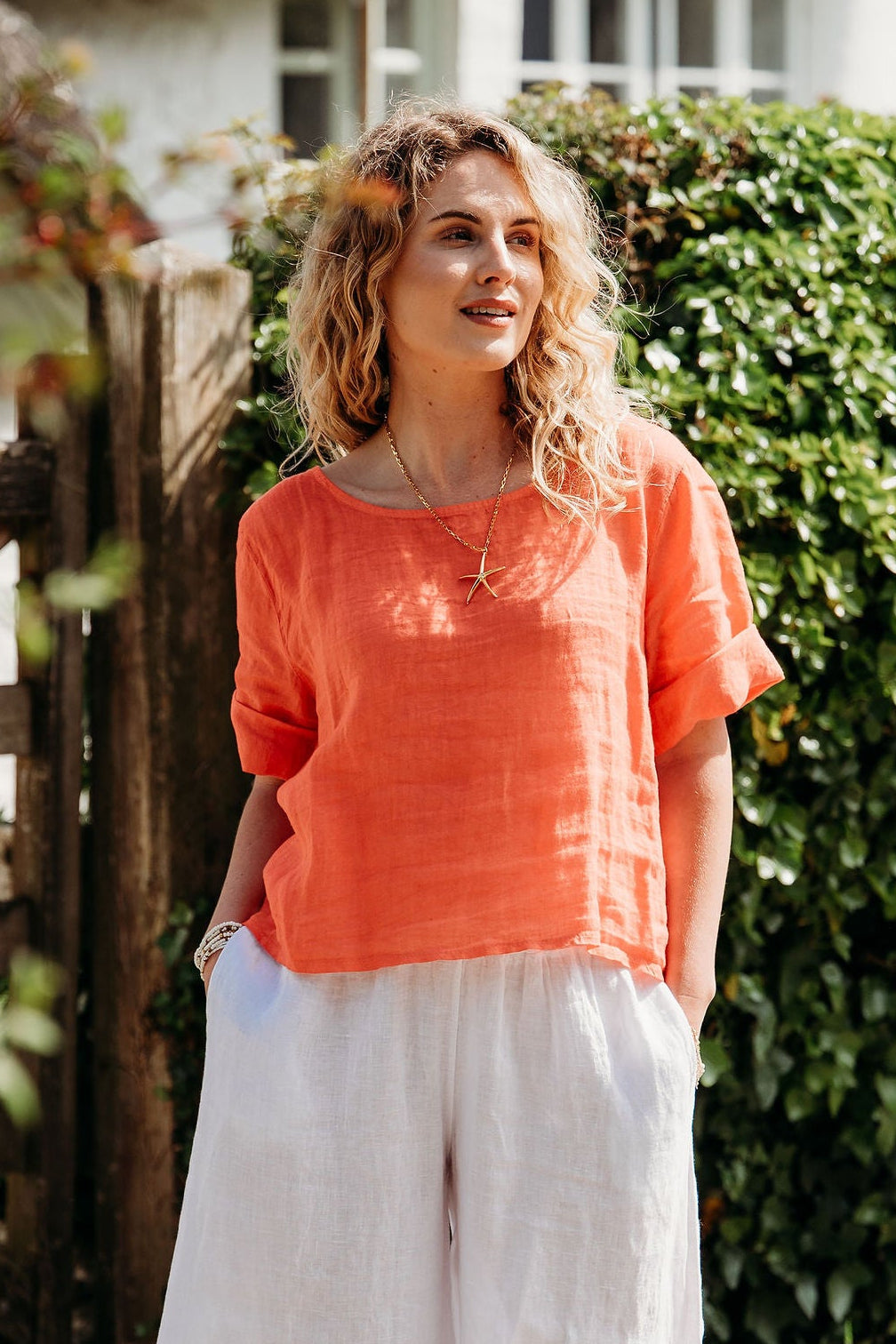 Woman in an orange top and white pants standing outdoors near a thatched-roof building.