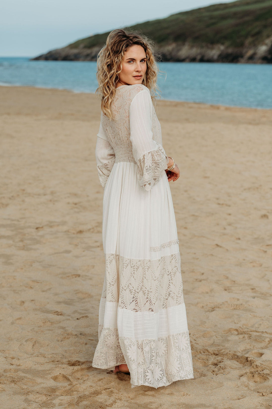 Woman in a long, light-colored dress standing on a sandy beach with ocean and cliffs in the background.