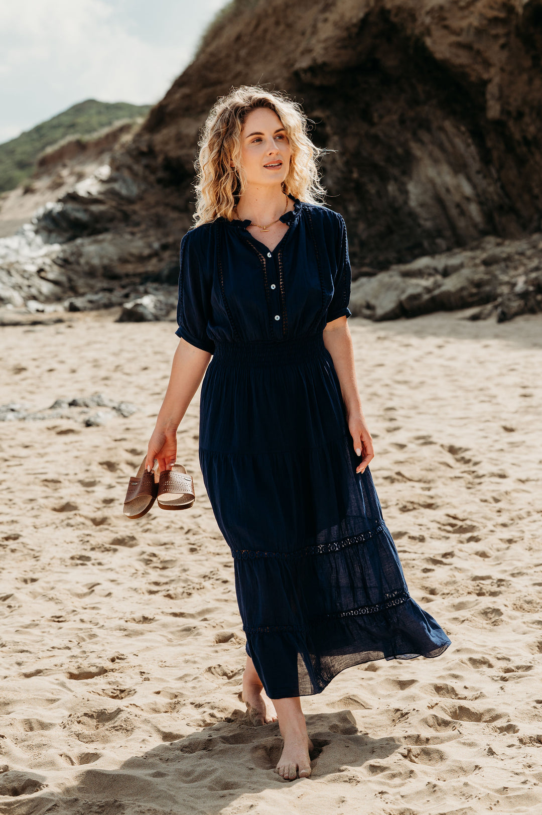 Woman in a dark blue dress standing on a sandy beach with rocky cliffs in the background