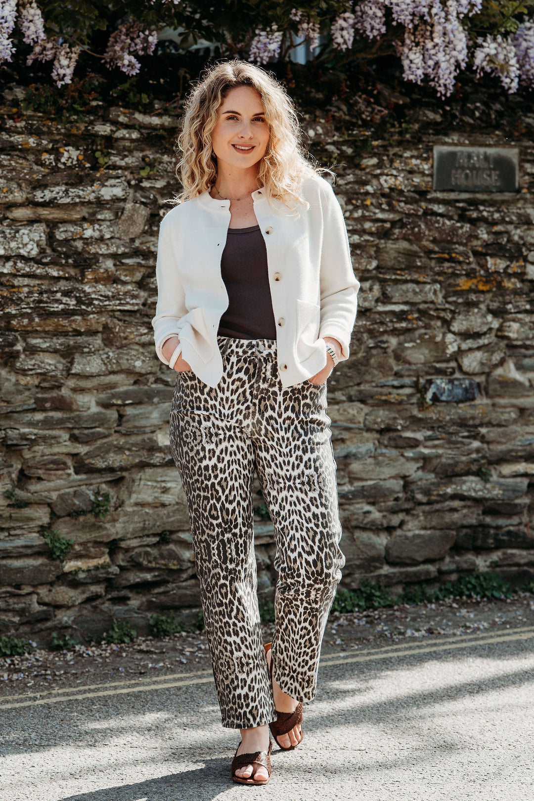 Woman in a white jacket and leopard print pants standing in front of a stone wall with wisteria.