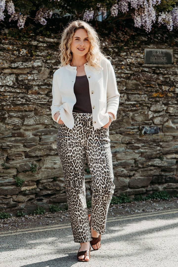 Woman in a white jacket and leopard print pants standing in front of a stone wall with wisteria.