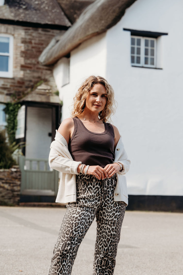 Woman standing in front of a white house wearing a brown tank top and leopard print pants.