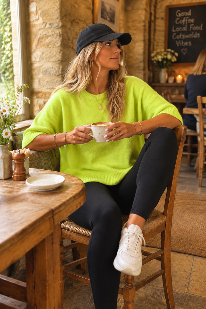 A woman sitting in a rustic cafe setting wearing a bright lime green knit sweater, black leggings, white sneakers, and a black baseball cap, holding a white coffee mug.