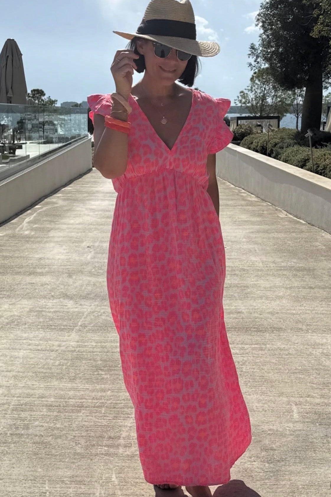 Woman in a pink dress and straw hat standing on a wooden deck with trees and sky in the background.