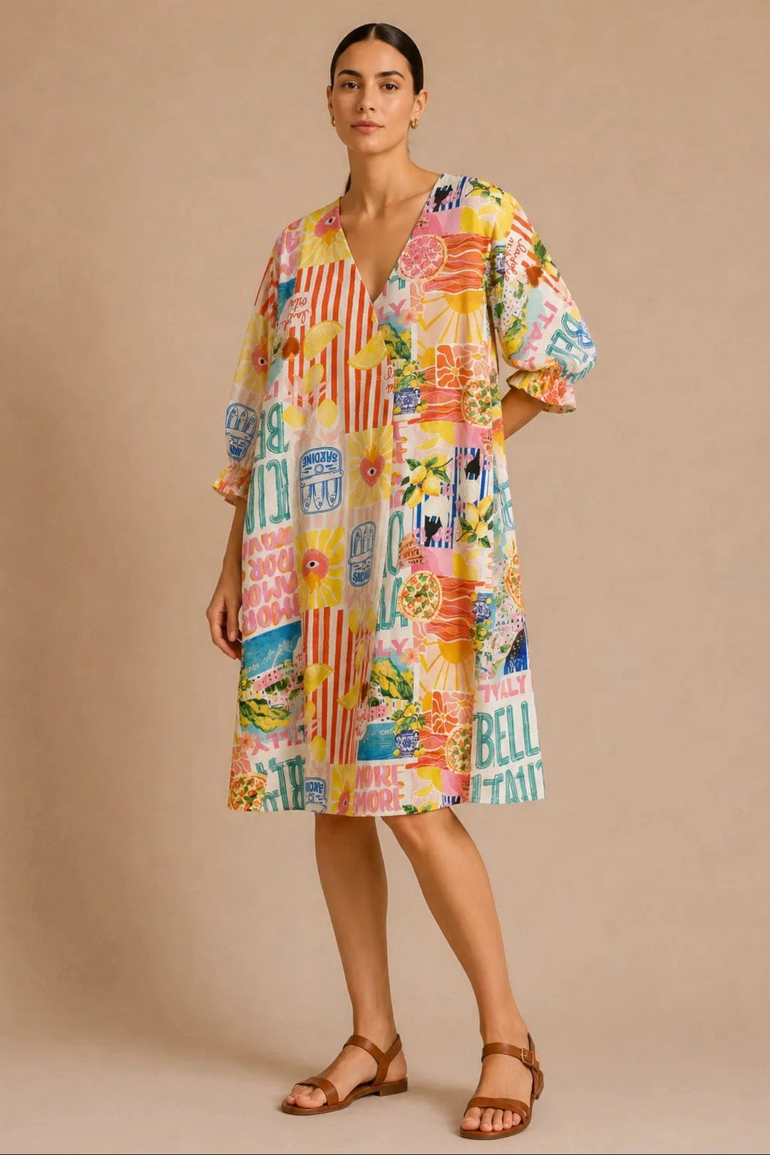 A woman modeling a short-sleeve, knee-length dress with a colorful collage print featuring retro travel signs, palm trees, and beach motifs, set against a solid beige studio background.