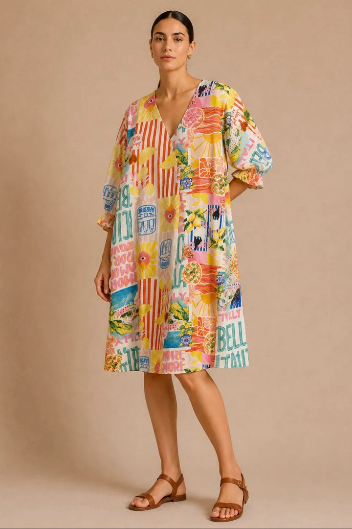 A woman modeling a short-sleeve, knee-length dress with a colorful collage print featuring retro travel signs, palm trees, and beach motifs, set against a solid beige studio background.