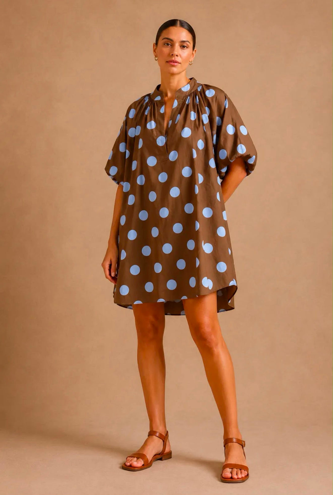 A woman modeling a brown short-sleeve mini dress with a light blue polka dot pattern, paired with brown leather sandals, against a solid beige studio backdrop.