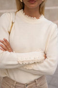 A woman wearing a classic cream-colored crew neck jumper with ruffle broderie pie crust trim on the neckline and cuffs.
