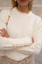 A woman wearing a classic cream-colored crew neck jumper with ruffle broderie pie crust trim on the neckline and cuffs.