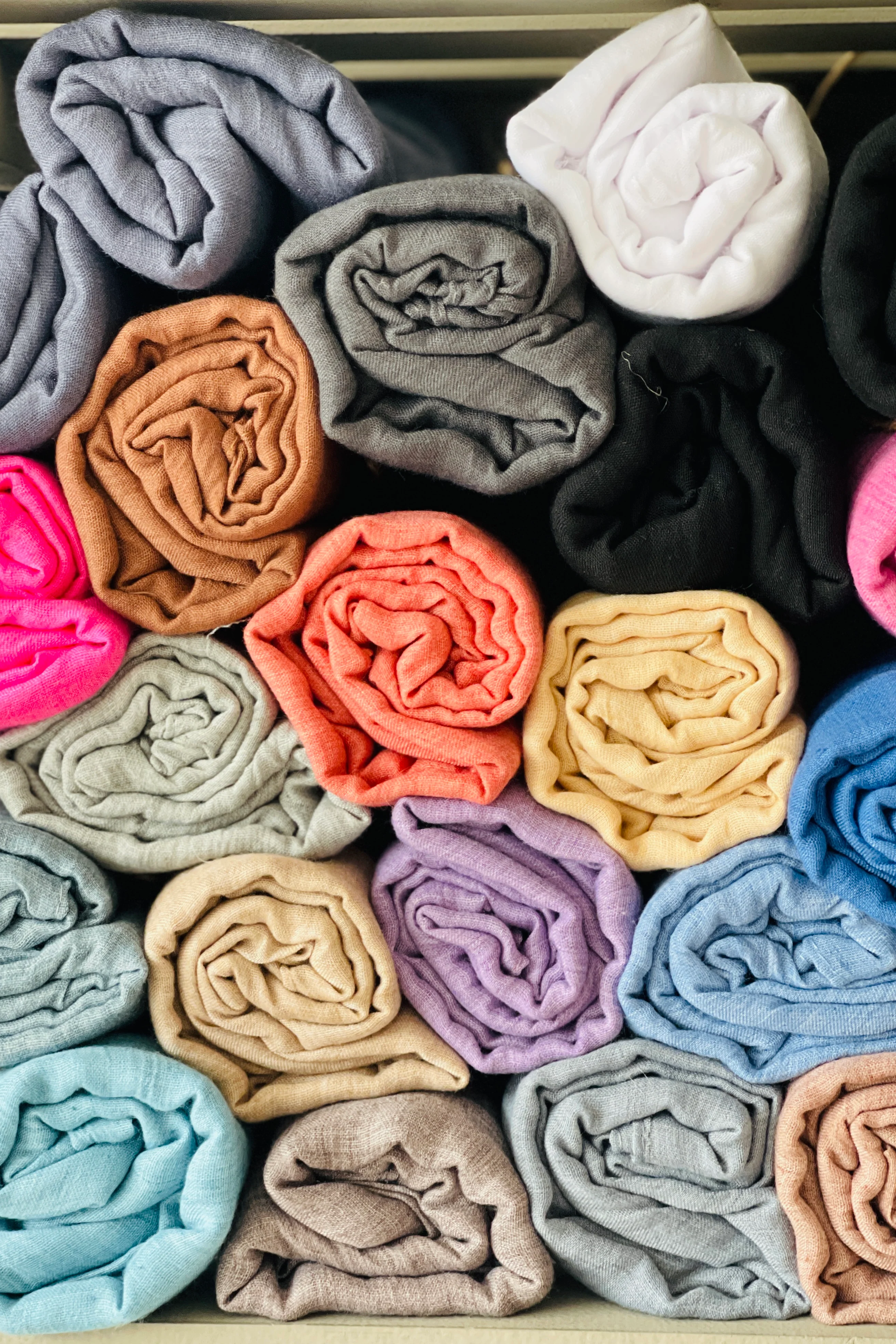 A collection of folded cotton scarves in various colors including gray, black, pink, orange, blue, and purple, arranged in a pile.