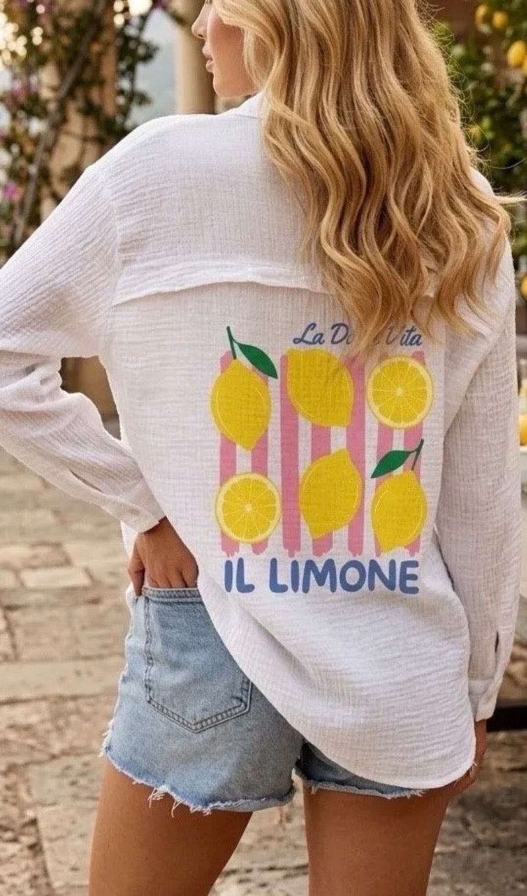 A woman wearing a white textured long-sleeve shirt with a colorful lemon graphic on the back, paired with denim shorts, photographed outdoors.