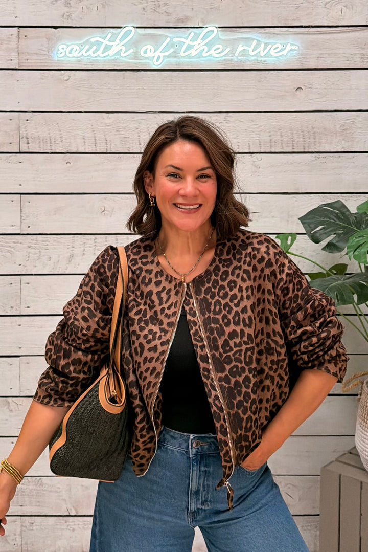 A woman posing in a leopard-print bomber jacket, black top, and wide-leg blue jeans, carrying a dark green shoulder bag. She is standing in front of a white wood-paneled wall with a glowing 'south of the river' sign and a potted plant.