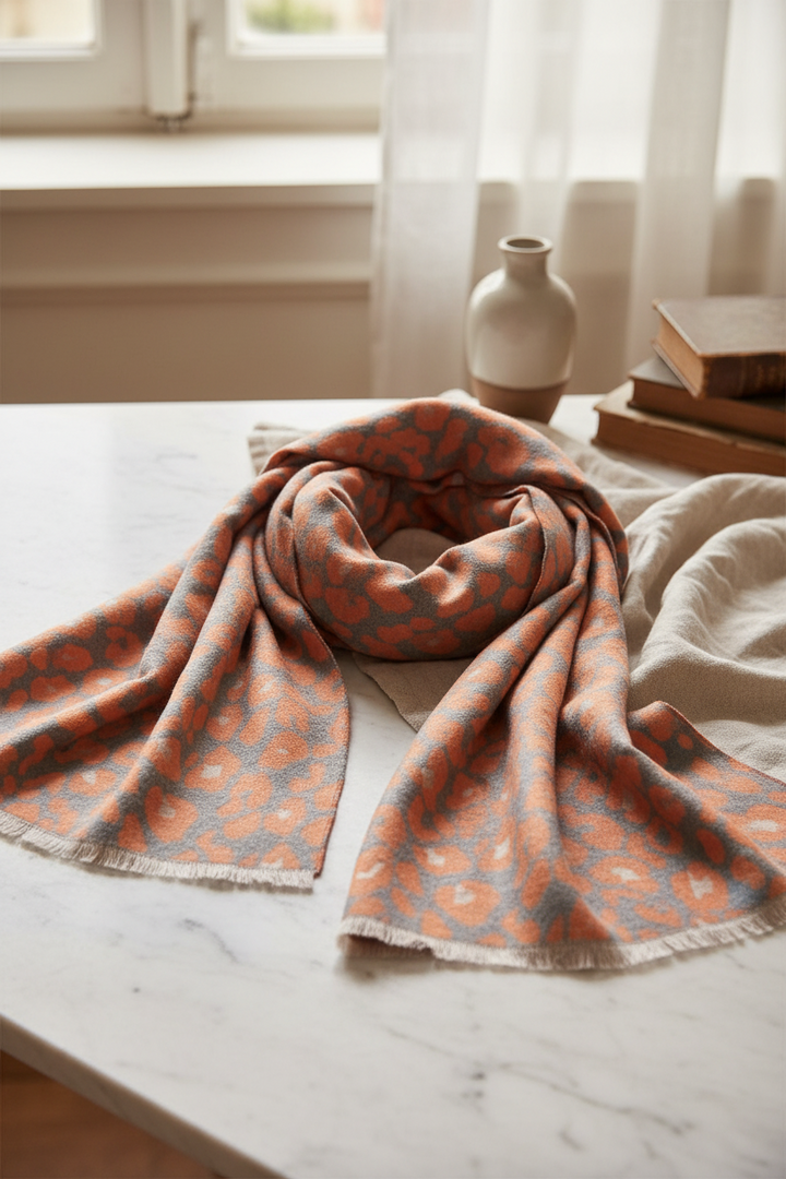 A lightweight woven scarf with a leopard print pattern in orange and gray, finished with a fringed edge, displayed on a clean white background.
