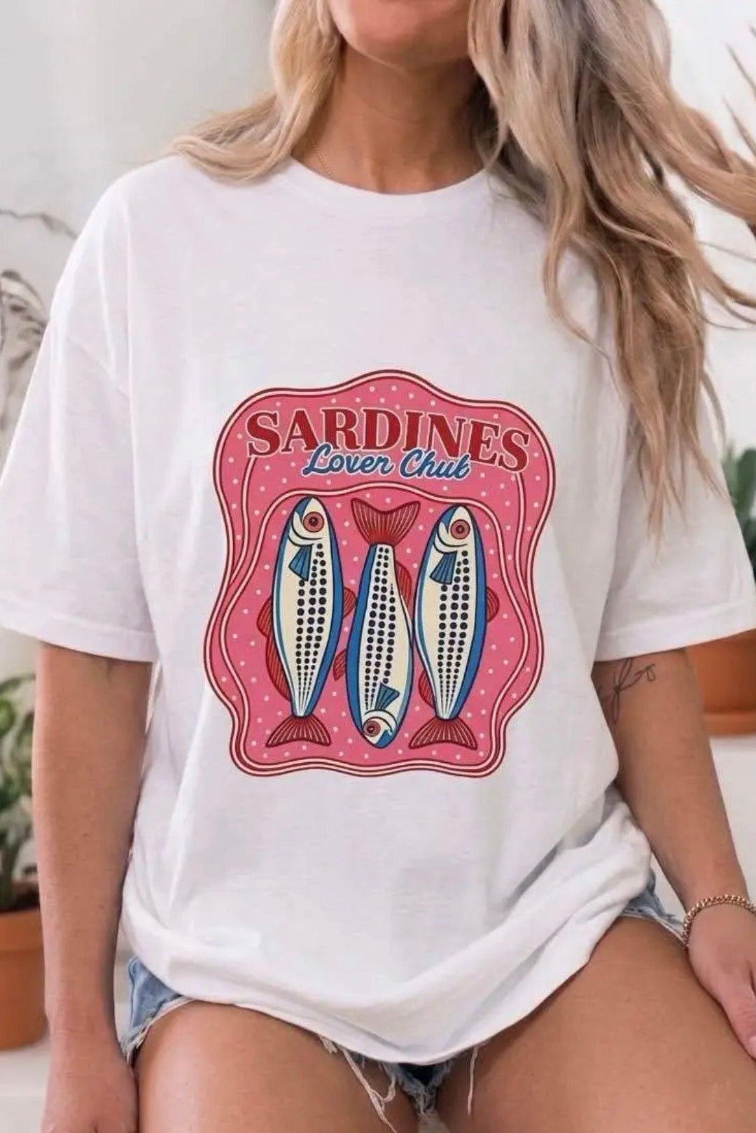 A woman wearing a white graphic t-shirt with a pink and blue 'Sardines Lover Club' design featuring three fish, paired with denim shorts in a brightly lit indoor setting.