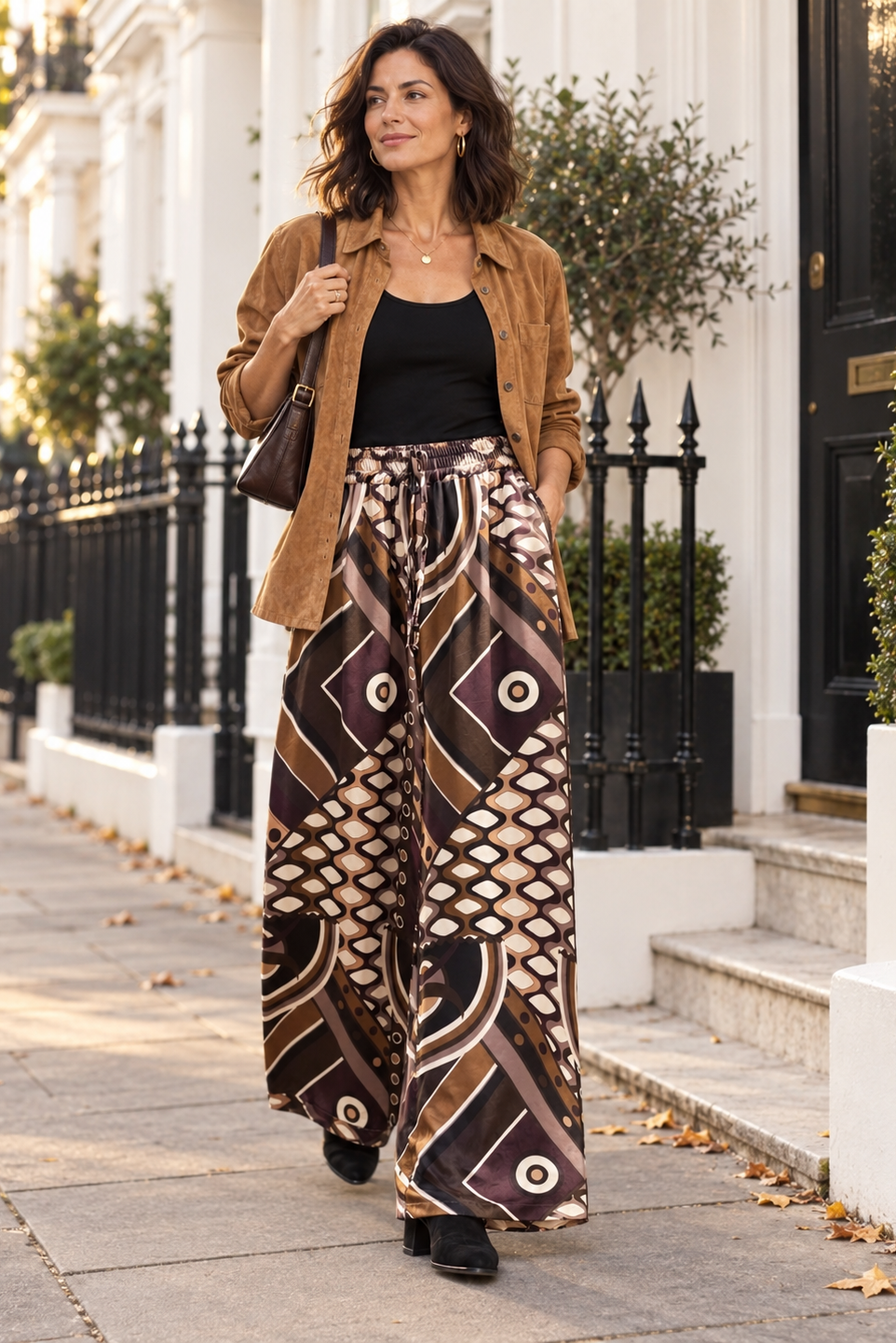 A woman walking on a city sidewalk wearing a brown suede jacket, black tank top, and wide-leg patterned pants in brown, beige, and black. She is carrying a brown shoulder bag.