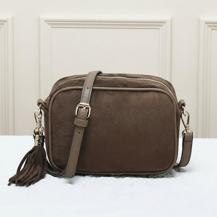 Brown leather crossbody bag with tassel on a white surface with a beige paneled wall background