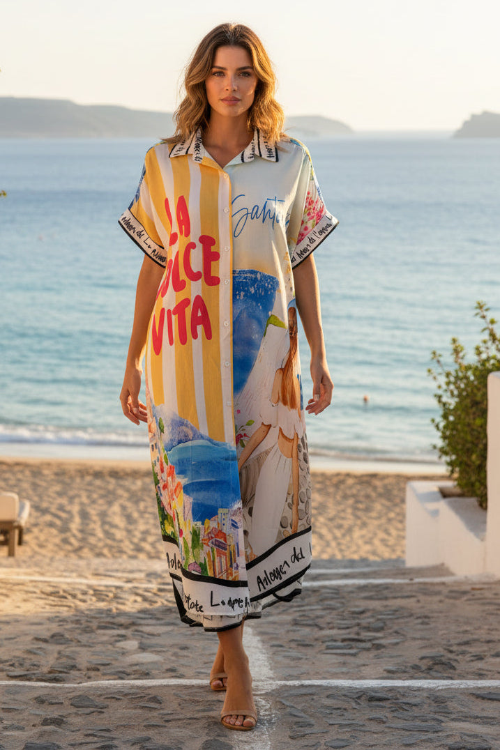 A woman wearing a short-sleeve kaftan dress with a colorful Mediterranean-themed print walks down a stone path towards the ocean, with white buildings and blue sea in the background.